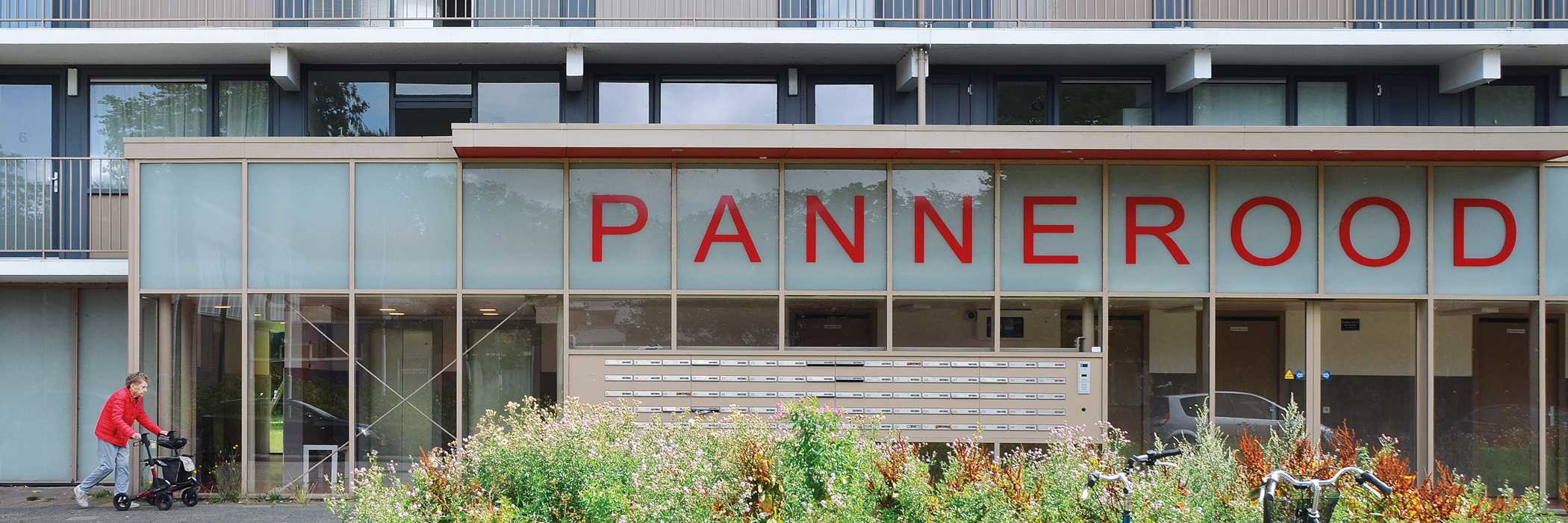 Een flatgebouw met beige kozijnen, voor het gebouw is een struik met kleurrijke planten.