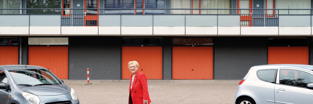 vooraanzicht van een flatgebouw met aan de onderkant garageboxen. op de foto staat een oudere dame in beeld