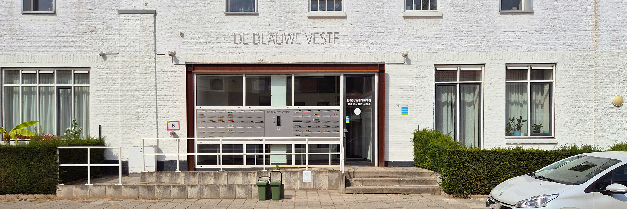 entree van een wit flatgebouw met parkeerplekken ervoor