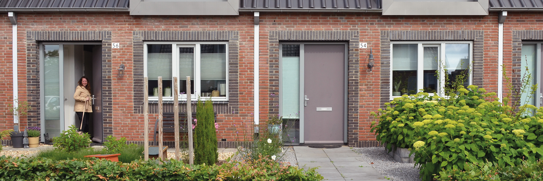 entree van twee woningen met een hele mooie groene tuin. de deuren zijn grijs en in de linker deur staat een mevrouw met een hond