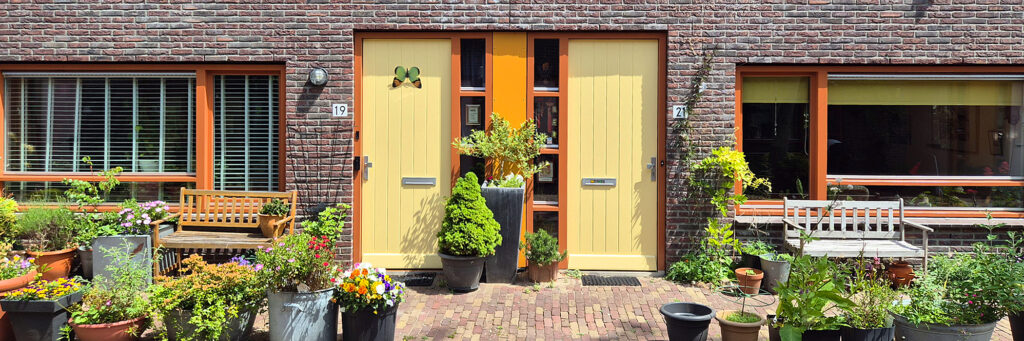Woningen met gele deuren en oranje kozijnen, de hele tuin staat vol met met bloempotten gevuld met kleurrijke bloemen