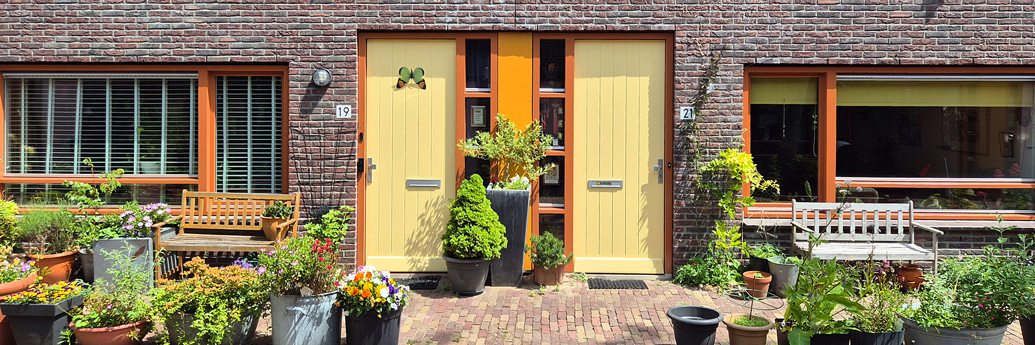 Woningen met gele deuren en oranje kozijnen, de hele tuin staat vol met met bloempotten gevuld met kleurrijke bloemen