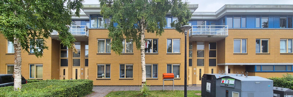 geel wooncomplex met een blauwe bovenverdieping. voor het complex staan bomen en groenstrook