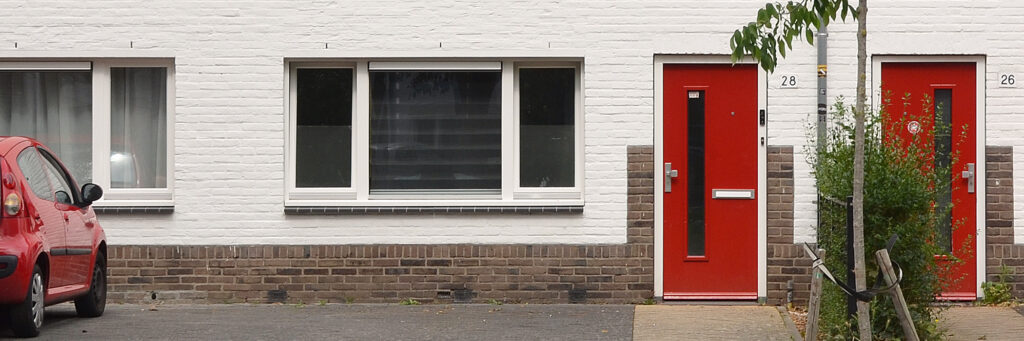 schakelwoningen met een rode deur en voor de rest alles wit. in de scheiding van de huizen staat een groen struikje